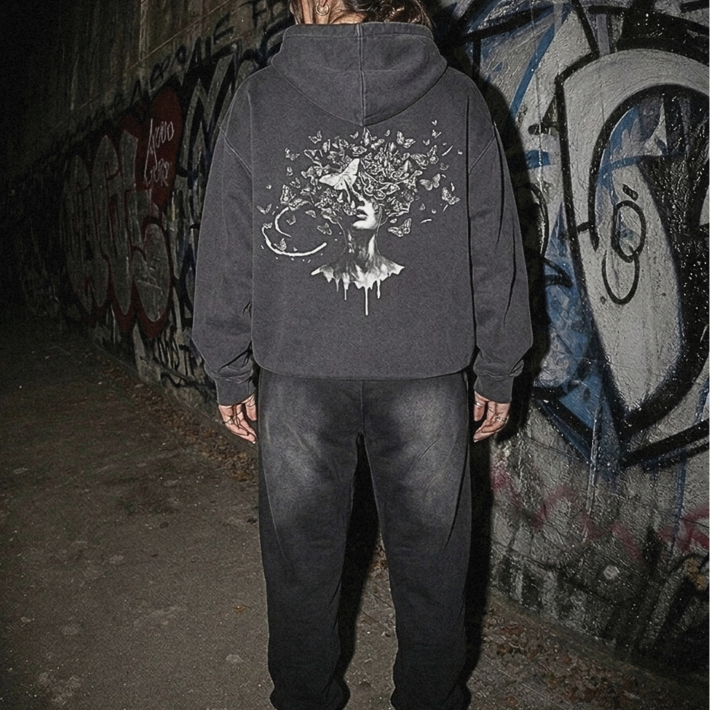A person viewed from behind wearing a dark gray hoodie with a large white graphic print on the back, paired with dark sweatpants and white sneakers, standing in front of a graffiti-covered wall.