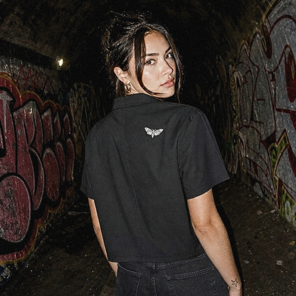 A woman stands in a dark graffiti-covered tunnel wearing a black short-sleeve crop top with a small white butterfly graphic on the upper back, paired with black jeans.