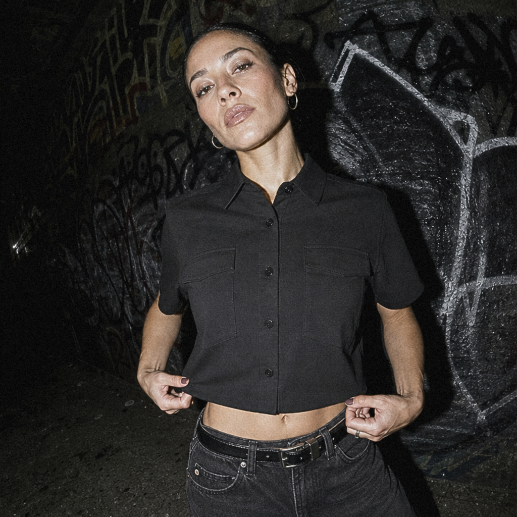 A woman posing in a black short-sleeve cropped button-up shirt and black jeans against a dark graffiti-covered wall.