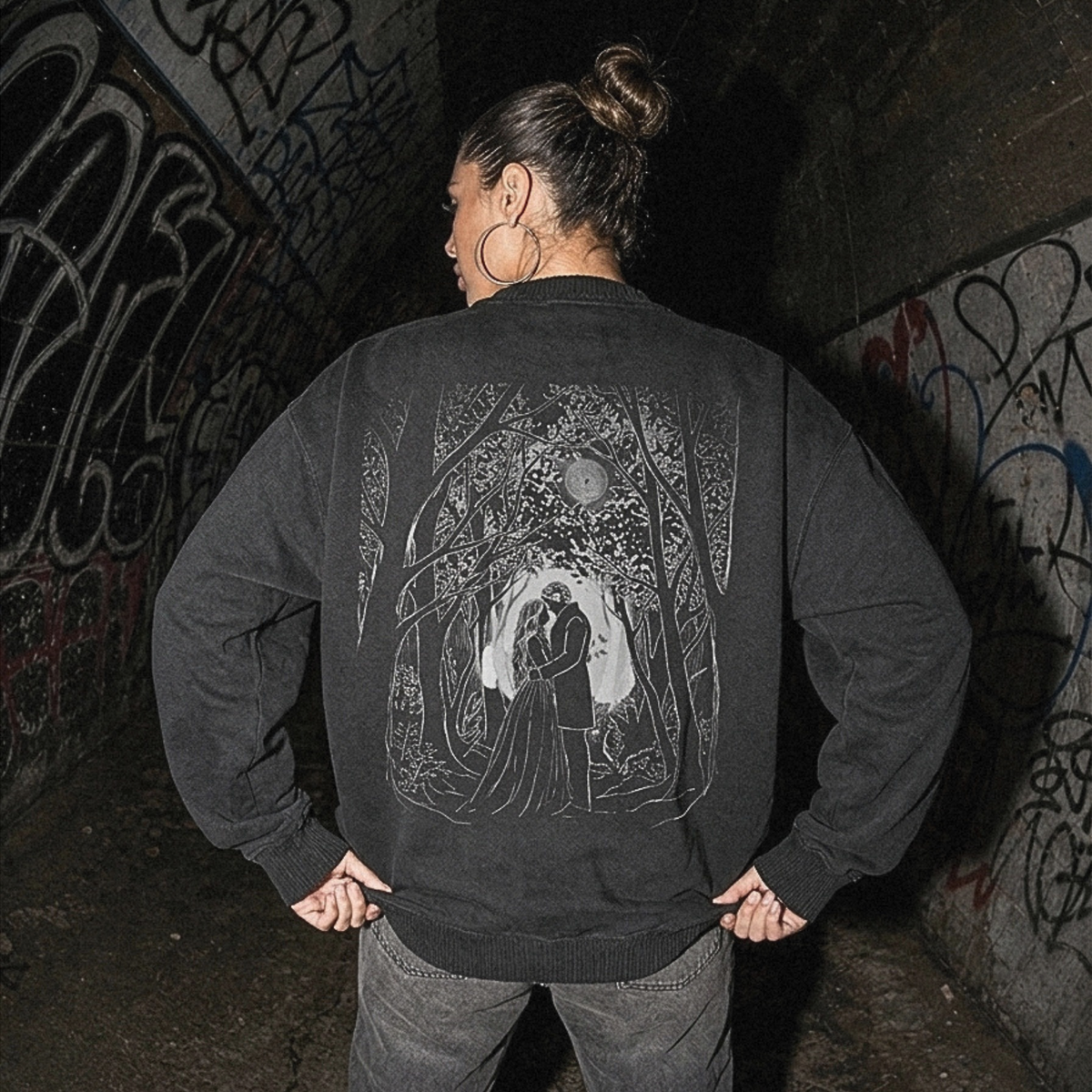 A person standing in a graffiti-covered tunnel, viewed from behind, wearing a dark charcoal gray sweatshirt with a large white graphic print of a cloaked figure on the back, paired with dark gray jeans.