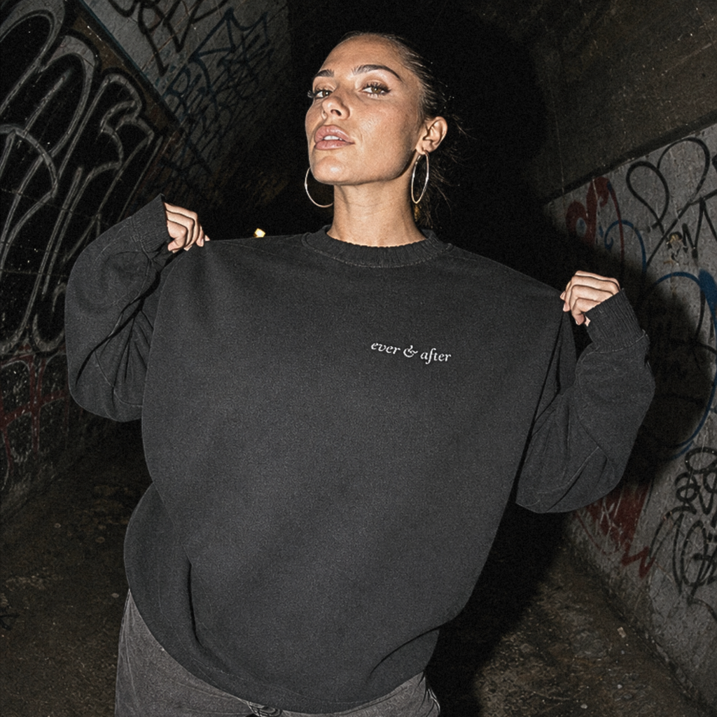 A woman posing in a dark, graffiti-covered tunnel wearing an oversized black crewneck sweatshirt with 'ever after' printed in white on the chest, paired with dark gray sweatpants.