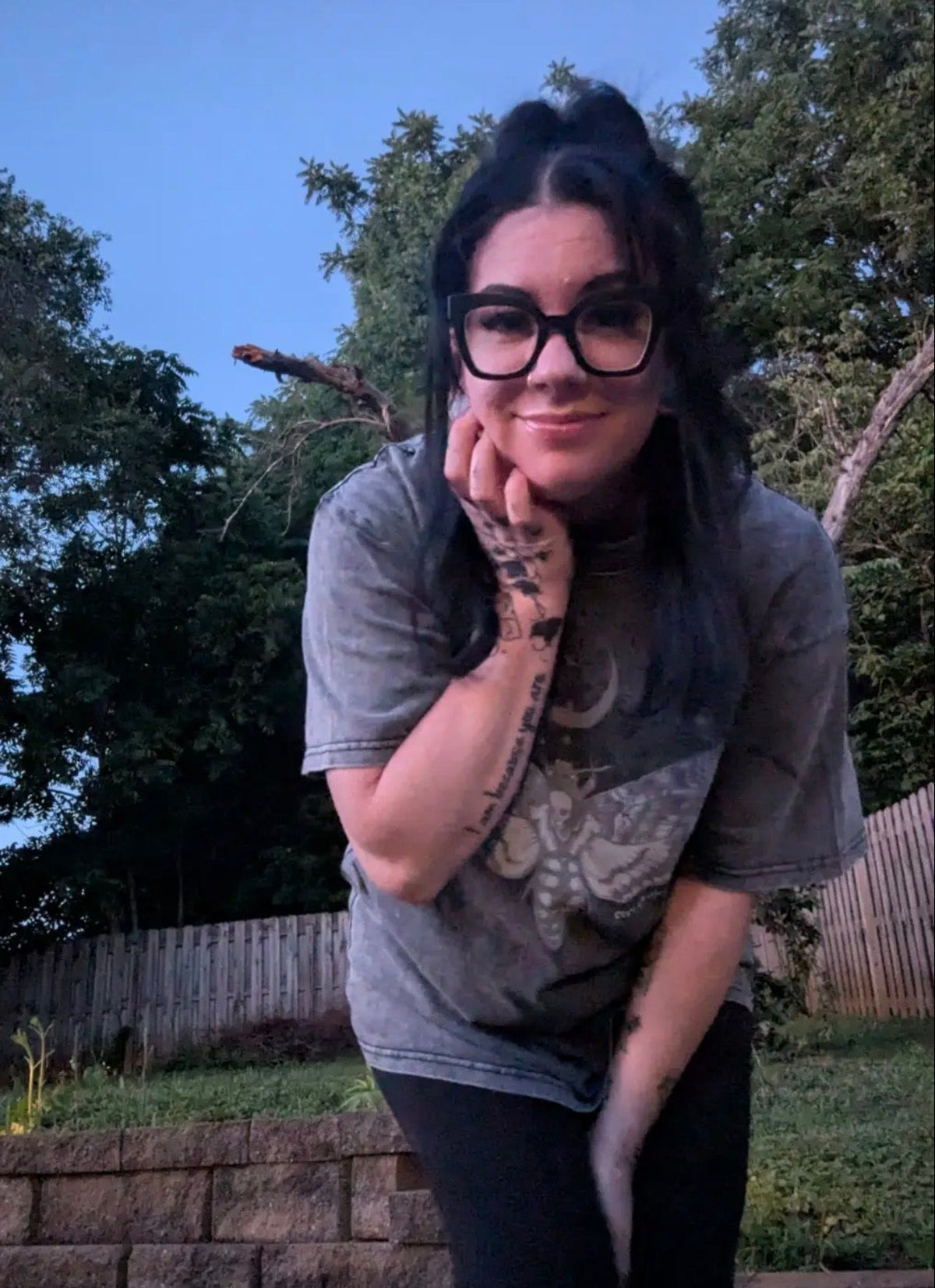 A woman with dark hair and black glasses posing outdoors in a gray graphic t-shirt and black pants, with trees and a wooden fence in the background.