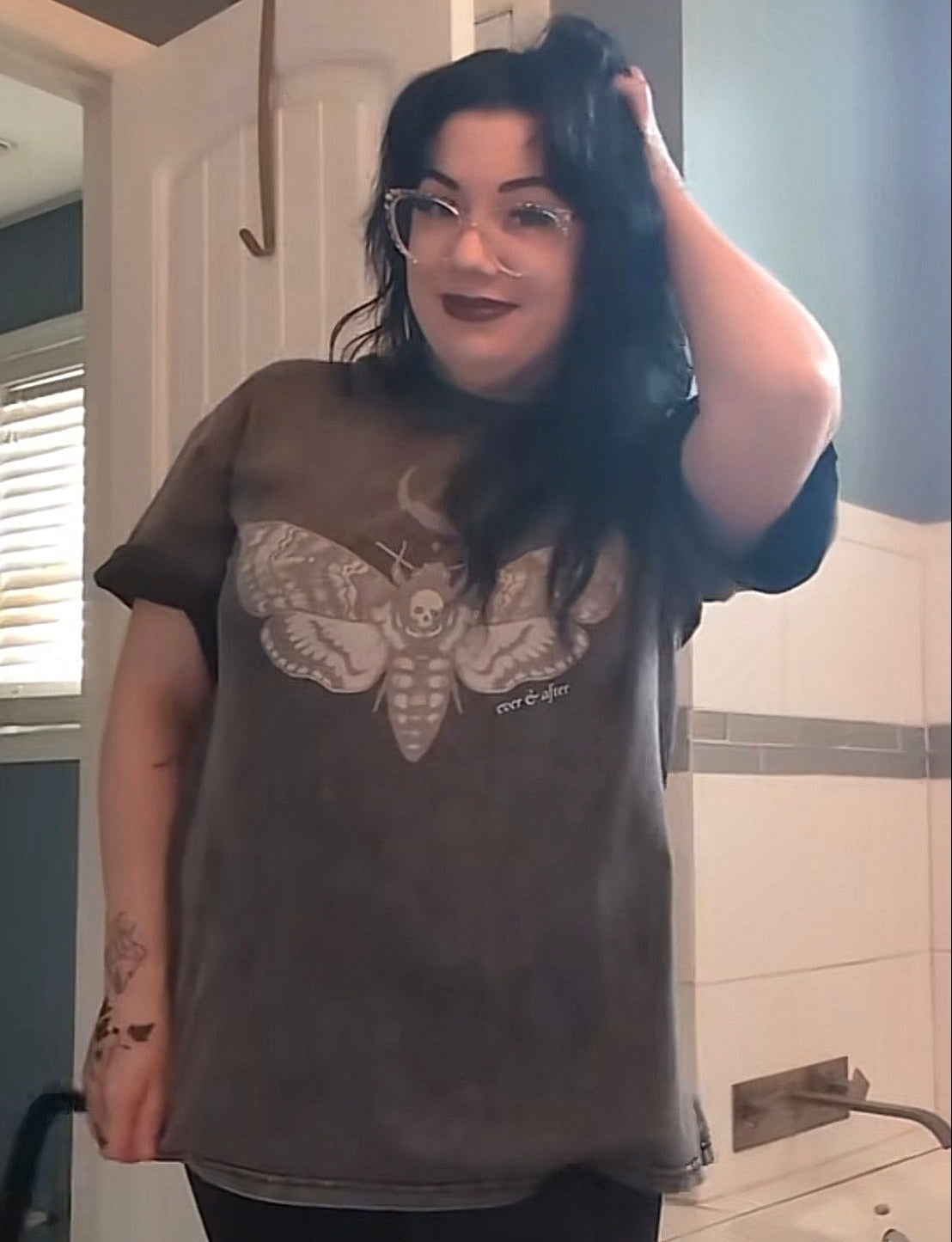 A woman with dark hair and glasses stands in a tiled bathroom wearing a faded brown graphic t-shirt with a large moth design on the front.