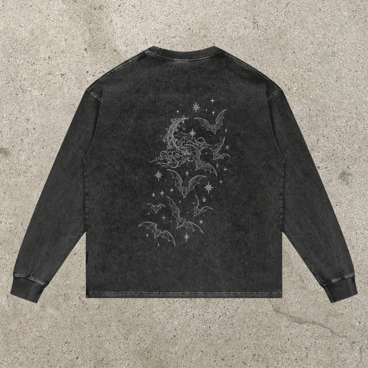 A dark gray mineral-wash long-sleeve shirt shown from the back, featuring a light gray celestial graphic of a crescent moon, stars, and bats, laid flat on a textured concrete surface.