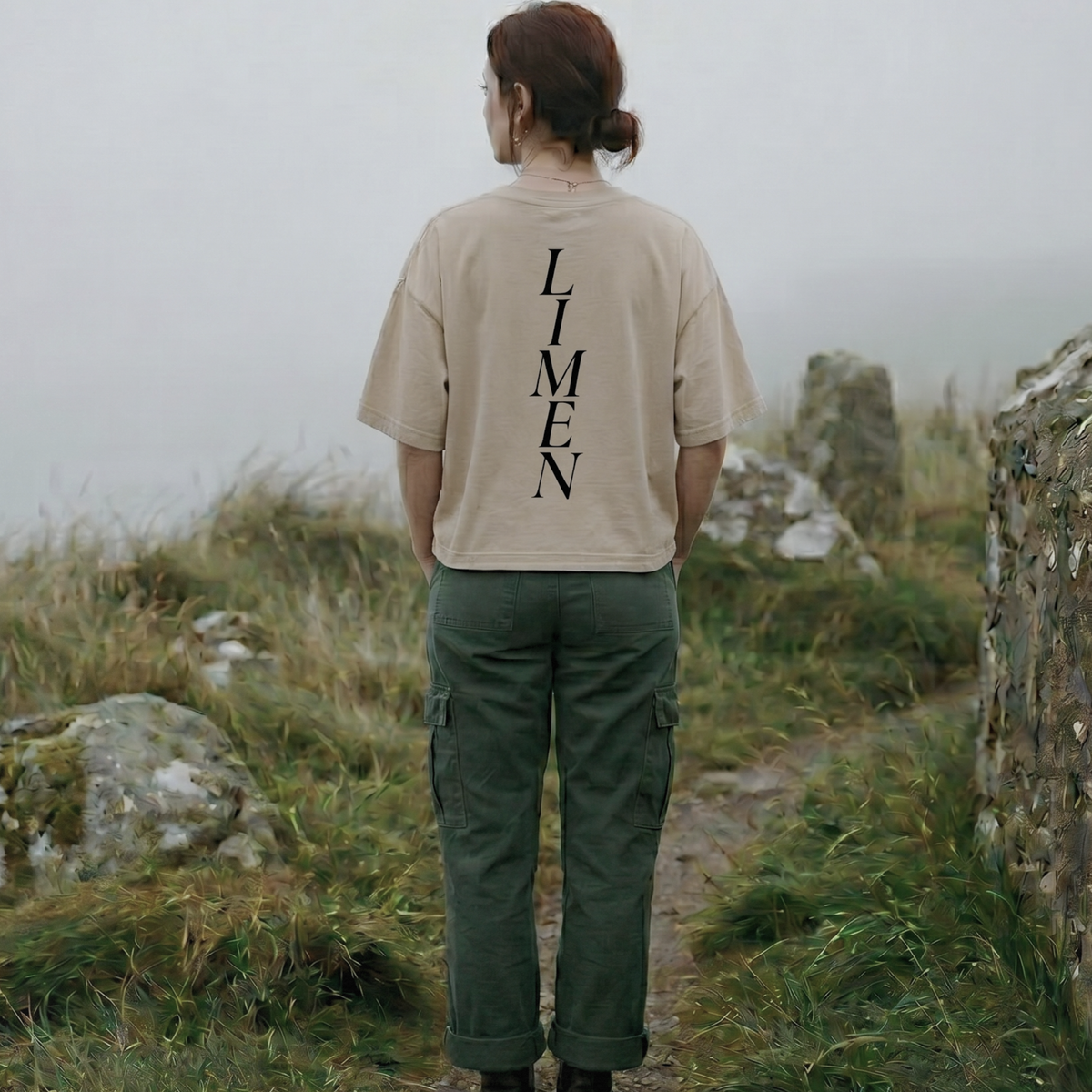 Person wearing a beige t-shirt with 'Limen' printed on the back, standing in a misty landscape.