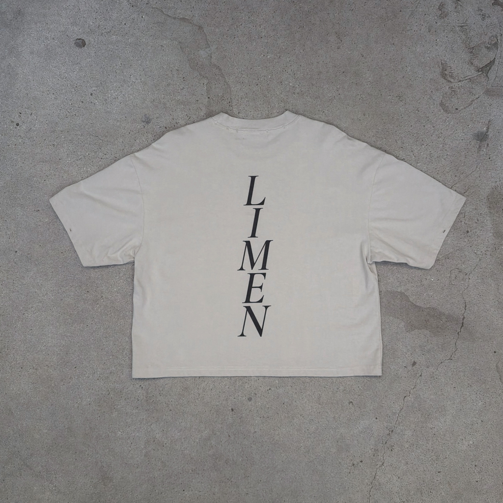 An oversized off-white t-shirt laid flat on a textured gray concrete surface, featuring the word 'LIMEN' printed vertically in black serif letters on the back.