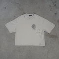 A distressed off-white cropped t-shirt with a circular graphic and text on the left chest, laid flat on a textured gray concrete surface.