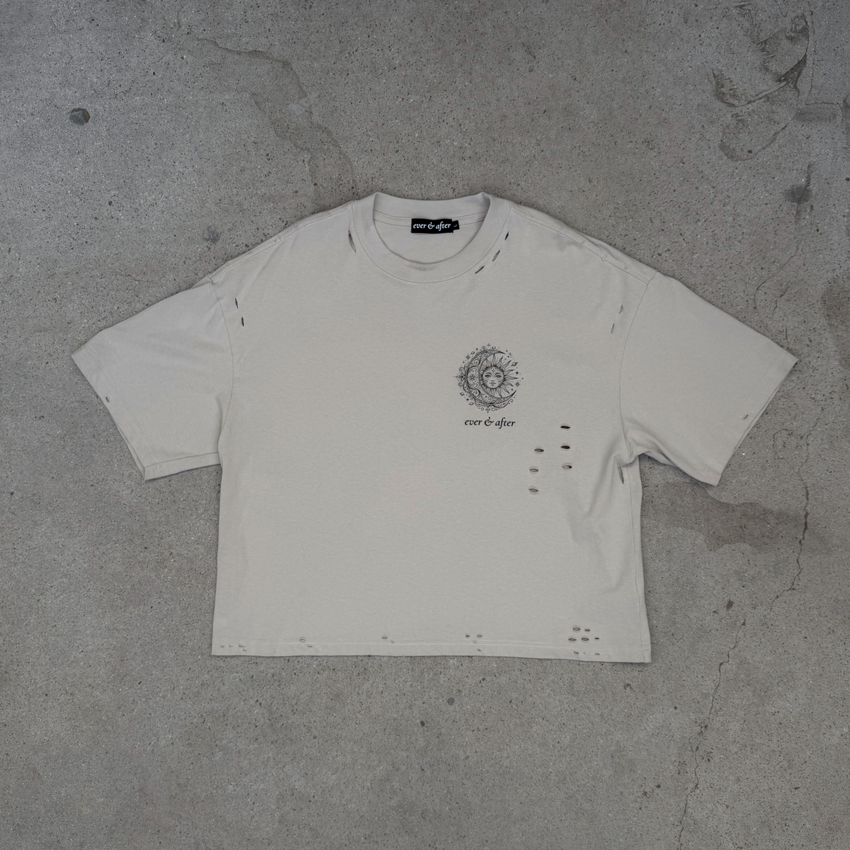 A distressed off-white cropped t-shirt with a circular graphic and text on the left chest, laid flat on a textured gray concrete surface.