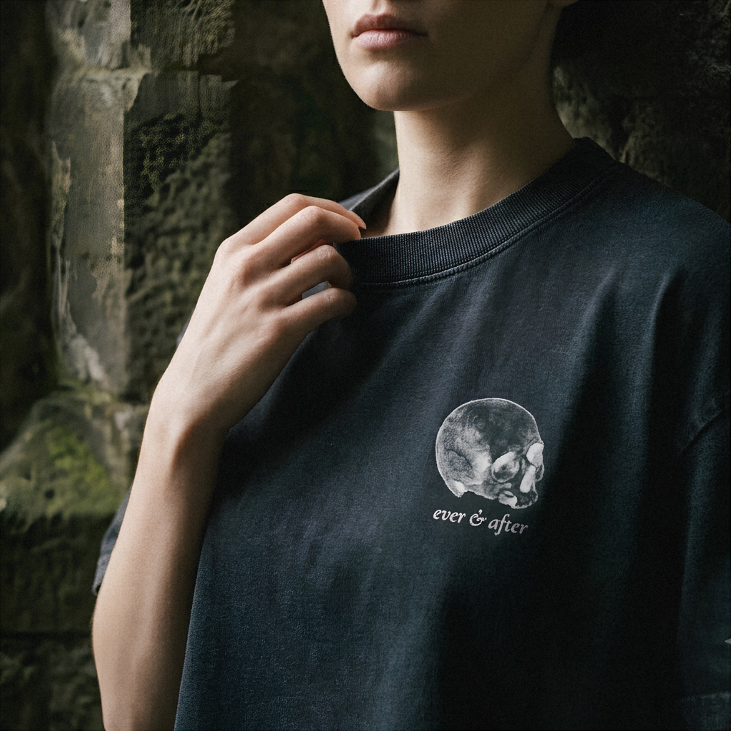 A close-up of a person wearing a dark charcoal crewneck t-shirt with a white skull graphic and the text 'ever & after' on the chest, set against a dark stone wall background.