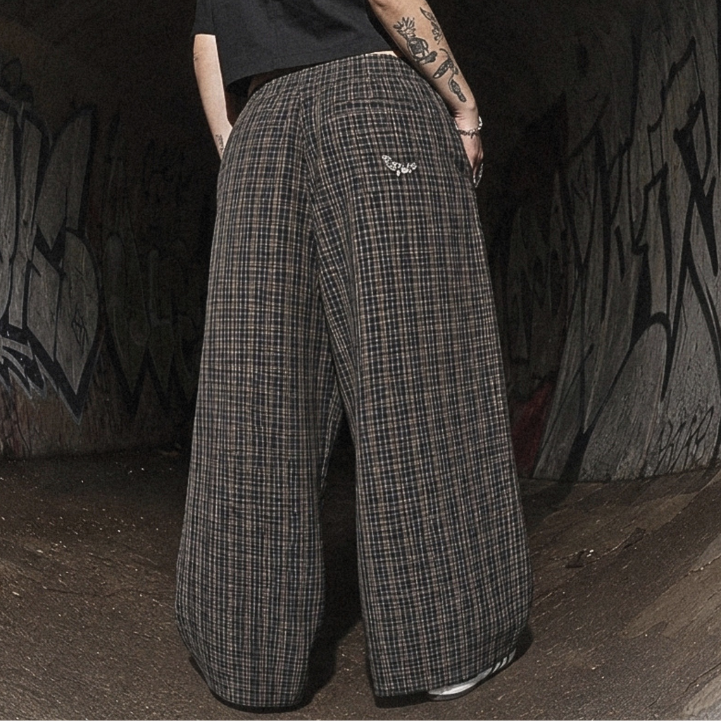A person standing in a dark graffiti-covered tunnel, viewed from behind, wearing a black short-sleeve shirt and very wide-leg plaid trousers in dark gray and black.
