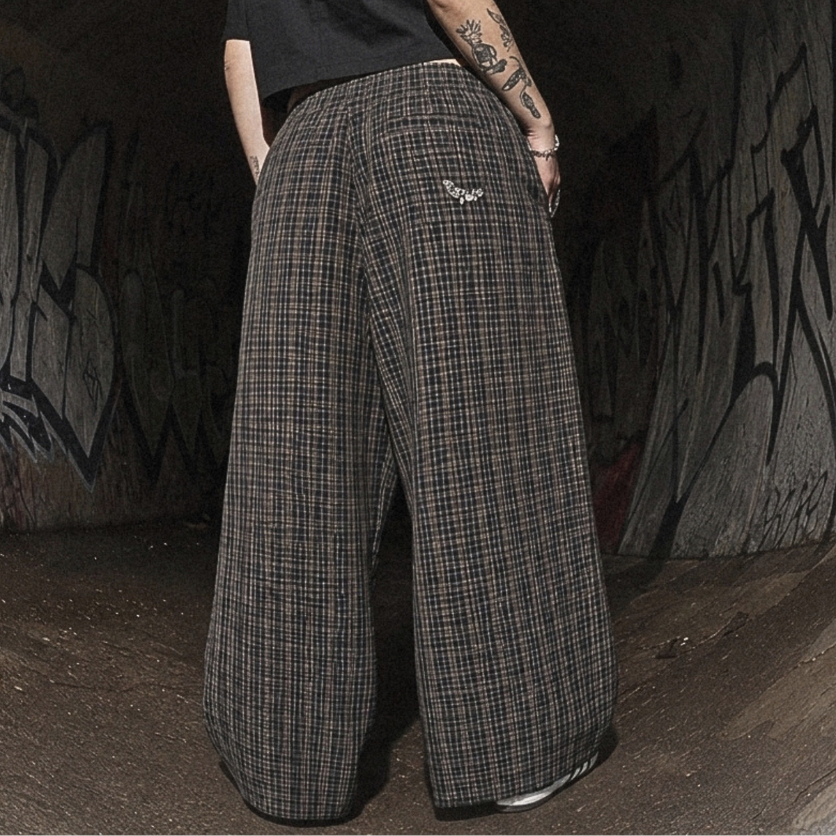A person standing in a dark graffiti-covered tunnel, viewed from behind, wearing a black short-sleeve shirt and very wide-leg plaid trousers in dark gray and black.