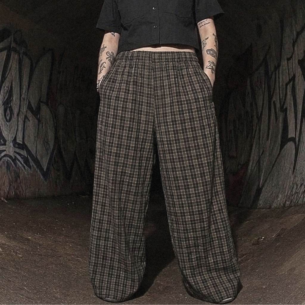 A person standing in a dark graffiti-covered tunnel wearing a black cropped short-sleeve button-up shirt and wide-leg plaid trousers.