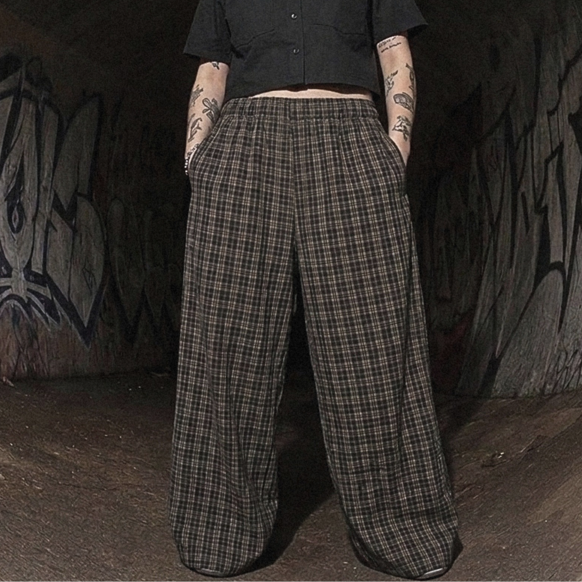 A person standing in a dark graffiti-covered tunnel wearing a black cropped short-sleeve button-up shirt and wide-leg plaid trousers.