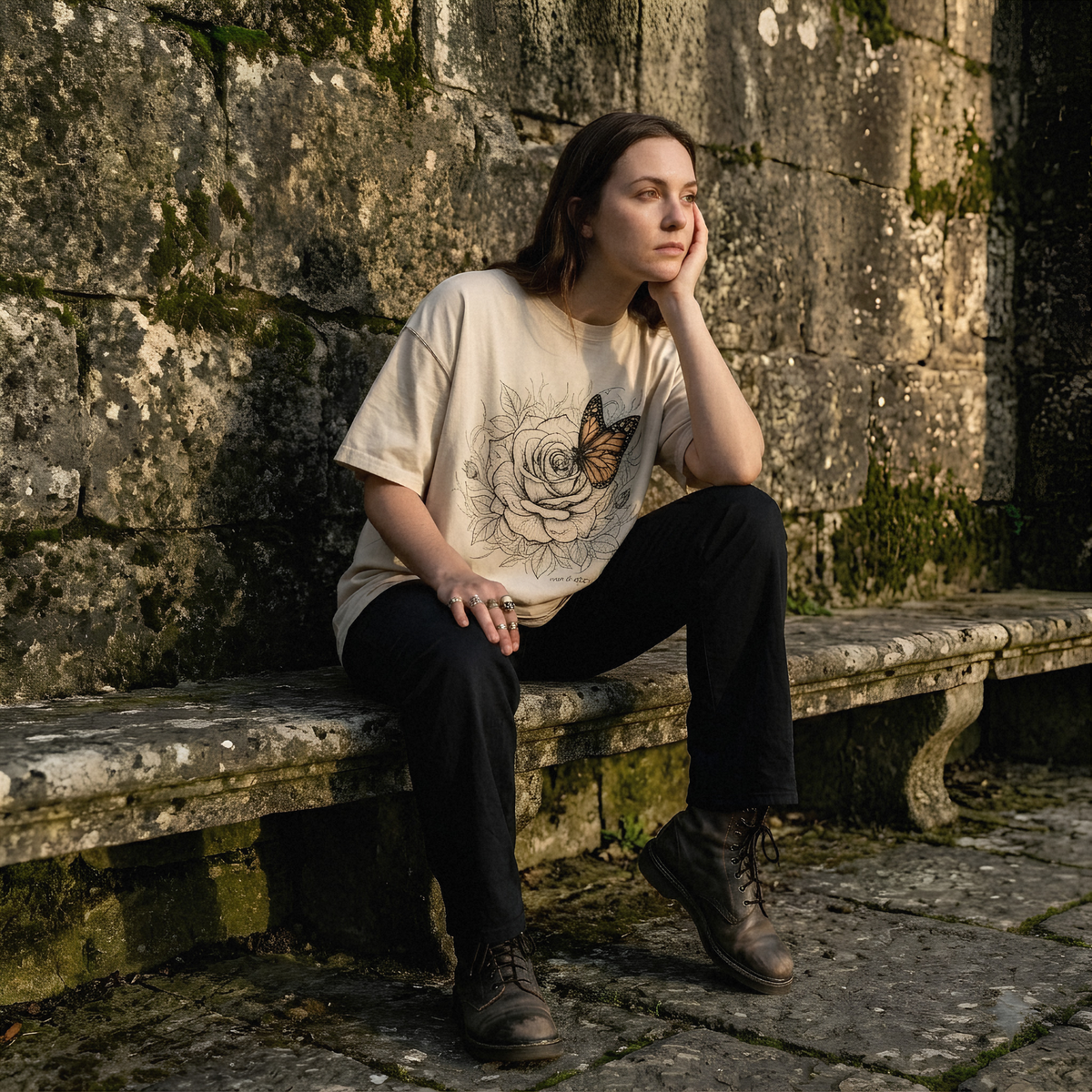 A woman sits on a weathered stone bench against a mossy stone wall, wearing a cream graphic t-shirt with a black rose and butterfly print, black pants, and brown lace-up boots.