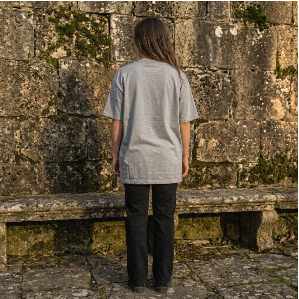 A person standing with their back to the camera, wearing a light gray heathered short-sleeve t-shirt and black pants, positioned in front of a mossy stone wall.