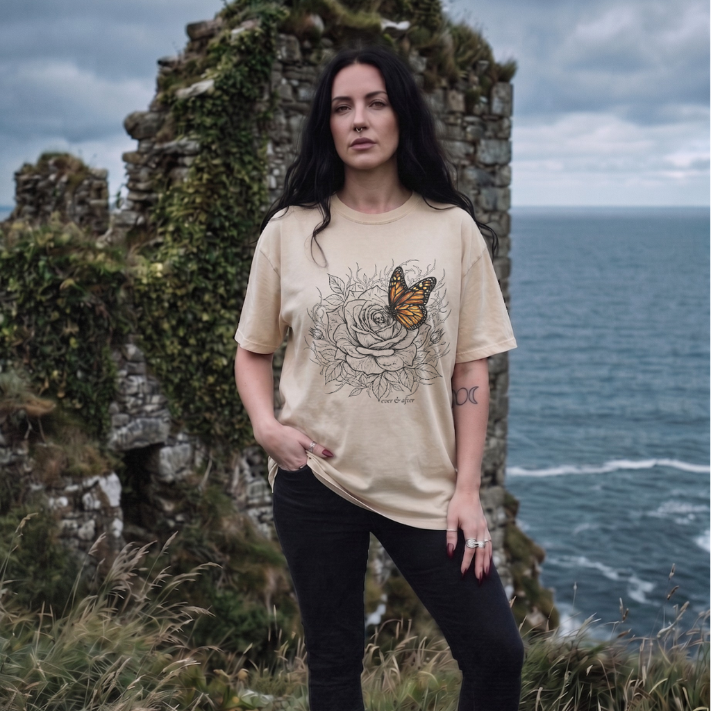 Woman wearing a beige t-shirt with a graphic design standing in front of a stone wall and ocean.