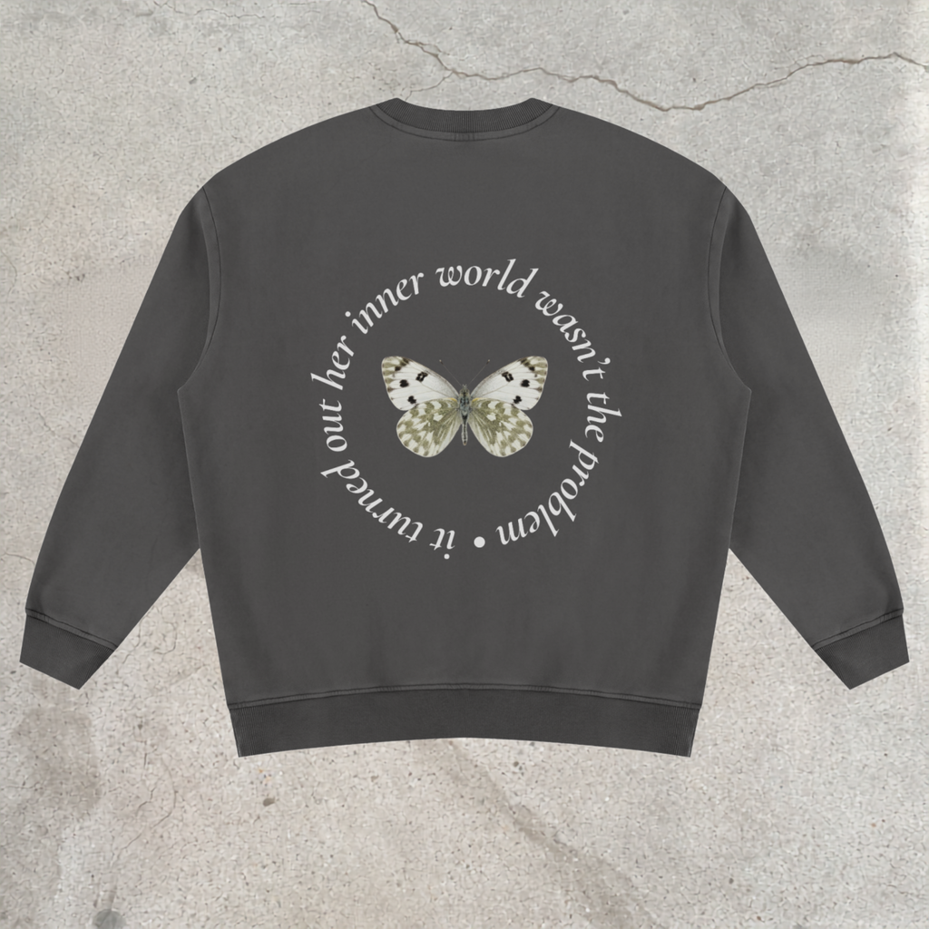 A dark gray crewneck sweatshirt shown from the back, featuring a circular white text graphic and a central butterfly design in white, black, and gold, laid flat on a textured concrete surface.