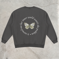 A dark gray crewneck sweatshirt shown from the back, featuring a circular white text graphic and a central butterfly design in white, black, and gold, laid flat on a textured concrete surface.