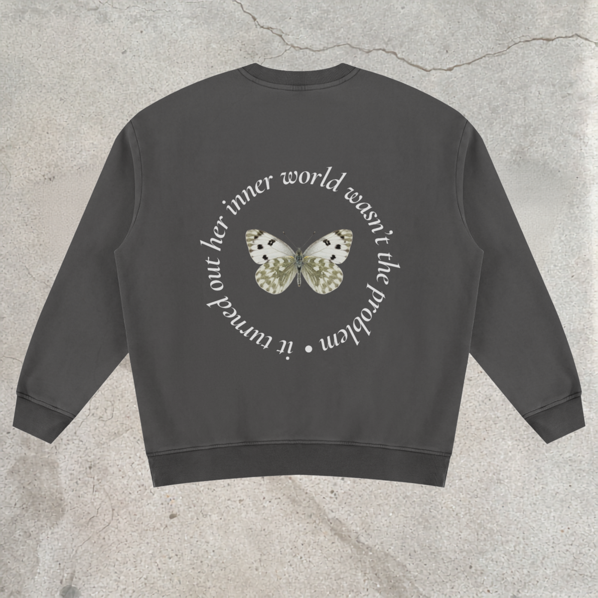 A dark gray crewneck sweatshirt shown from the back, featuring a circular white text graphic and a central butterfly design in white, black, and gold, laid flat on a textured concrete surface.