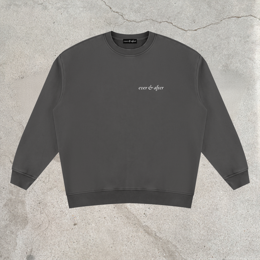 A dark gray crewneck sweatshirt with 'ever & after' printed in white script on the left chest, laid flat on a textured concrete background.