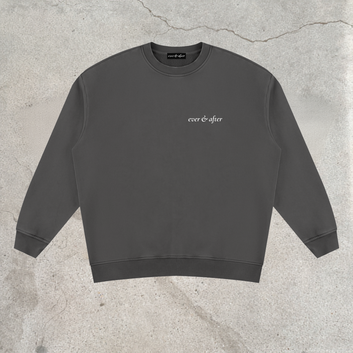 A dark gray crewneck sweatshirt with 'ever & after' printed in white script on the left chest, laid flat on a textured concrete background.