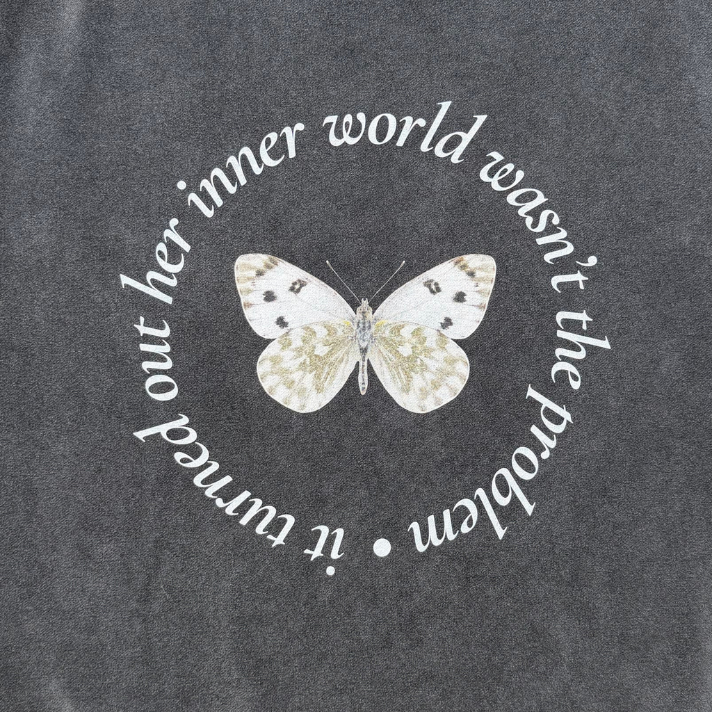 A close-up of a dark heather gray t-shirt featuring a white butterfly graphic in the center, surrounded by the white cursive text 'it turned out her inner world wasn't the problem'.