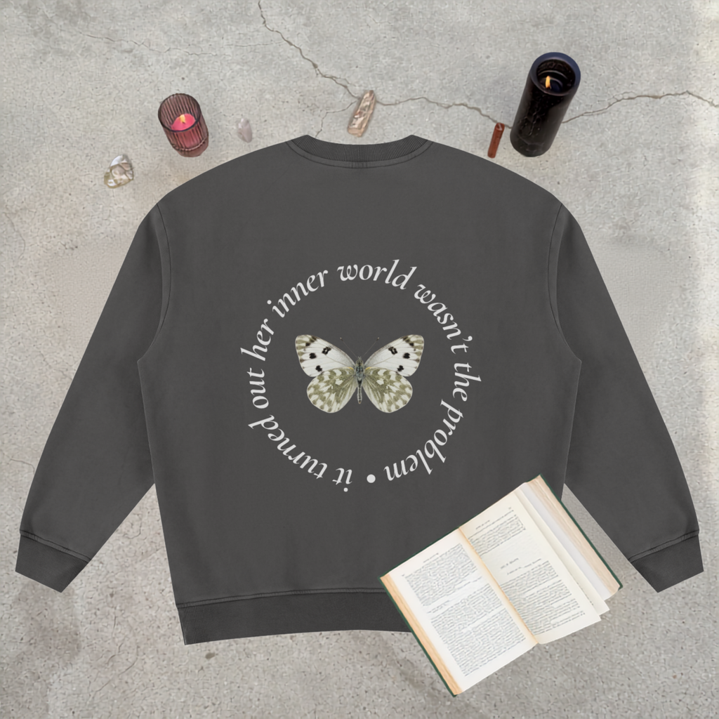 A dark charcoal gray crewneck sweatshirt laid flat on a textured concrete surface, featuring a graphic of a butterfly in the center with white and black spots, surrounded by a circular quote in white text. The sweatshirt is styled with an open book, crystals, and candles.