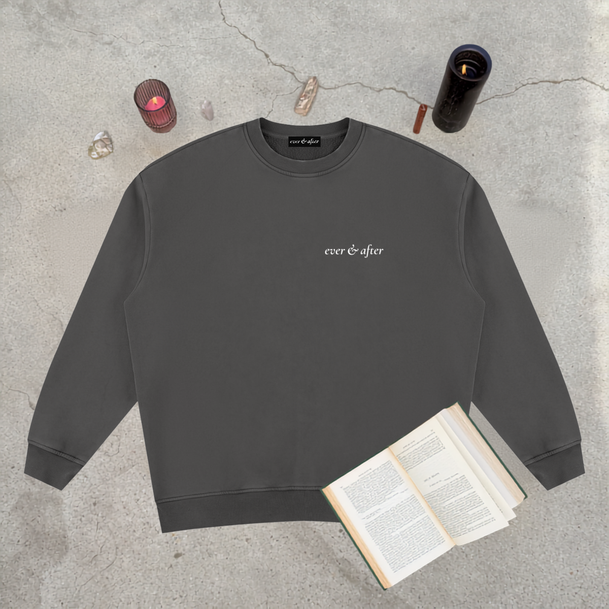 A dark gray crewneck sweatshirt with 'ever & after' printed in white script on the chest, styled in a flat lay on a concrete surface with an open book, crystals, and candles.