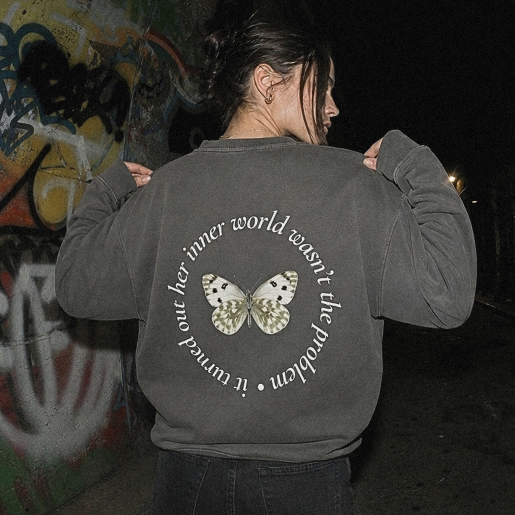 A person viewed from behind wearing a dark gray sweatshirt with a circular graphic on the back featuring a butterfly and the text 'it turned out her inner world wasn't the problem', set against a dark outdoor background with graffiti.