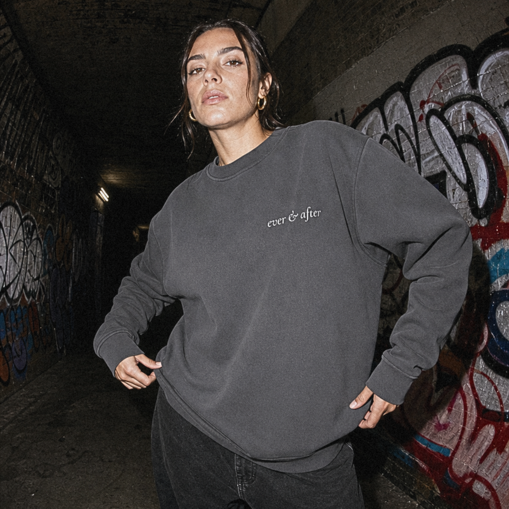 A woman wearing a dark charcoal gray crewneck sweatshirt with 'ever & often.' printed in white on the chest, standing in a graffiti-covered tunnel.