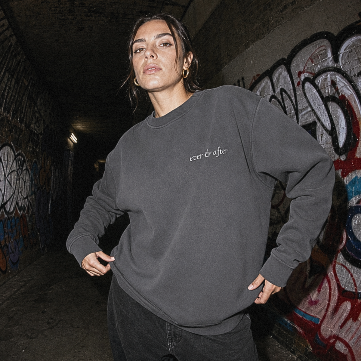 A woman wearing a dark charcoal gray crewneck sweatshirt with 'ever & often.' printed in white on the chest, standing in a graffiti-covered tunnel.