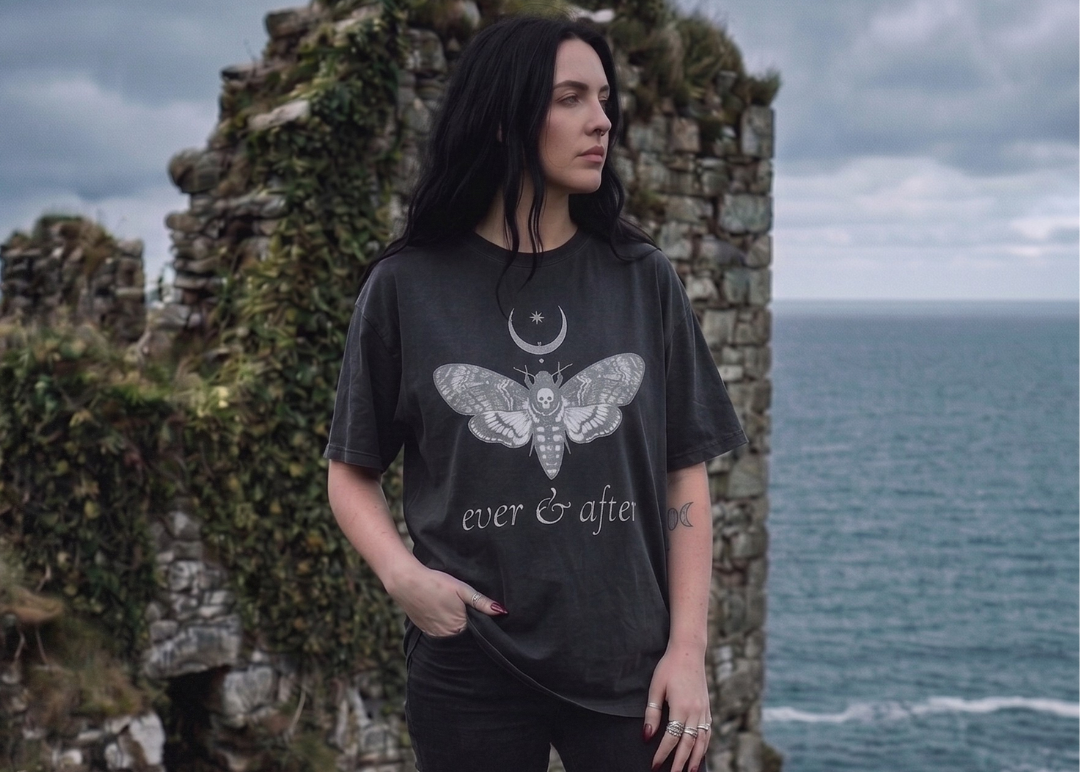 Person wearing a black t-shirt with a moth design and 'ever & after' text, standing in front of ocean cliffs.