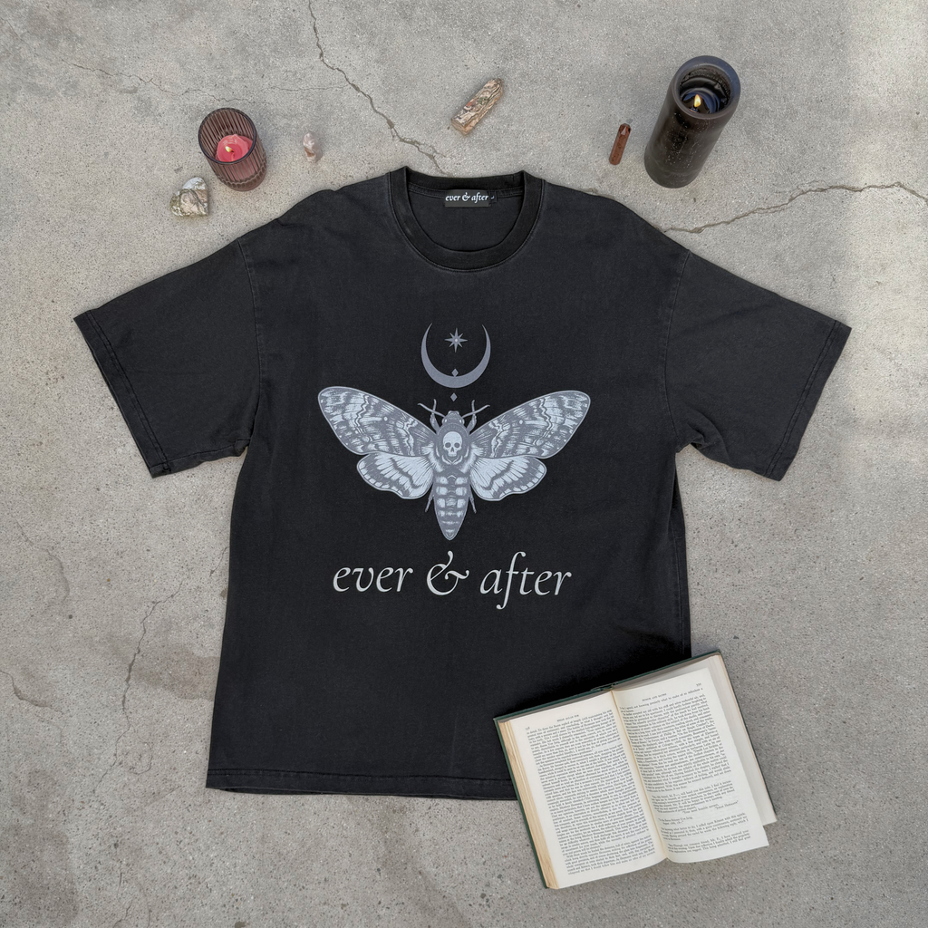 Black t-shirt with a moth and moon design on a concrete surface with candles and an open book.
