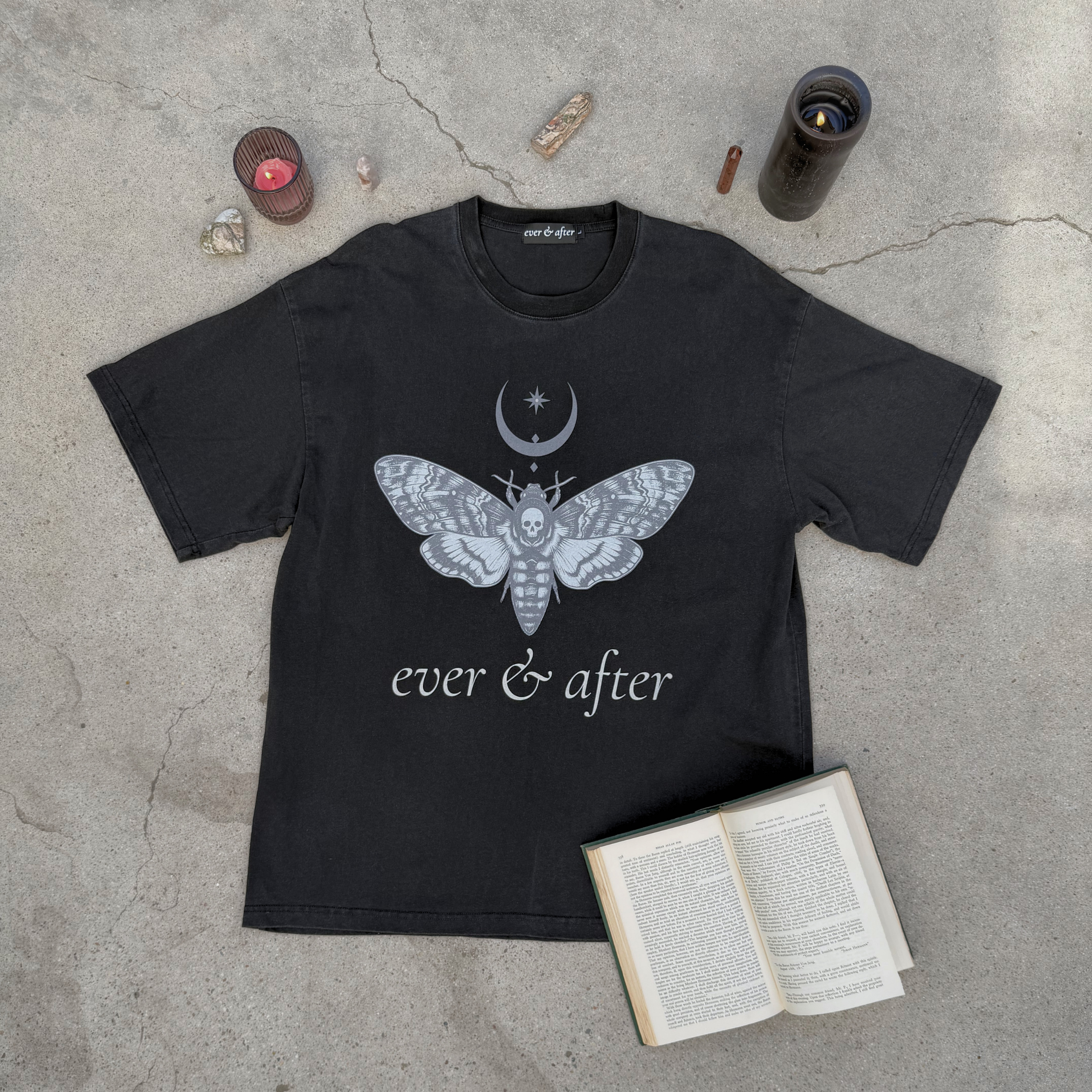 Black t-shirt with a moth and moon design on a concrete surface with candles and an open book.