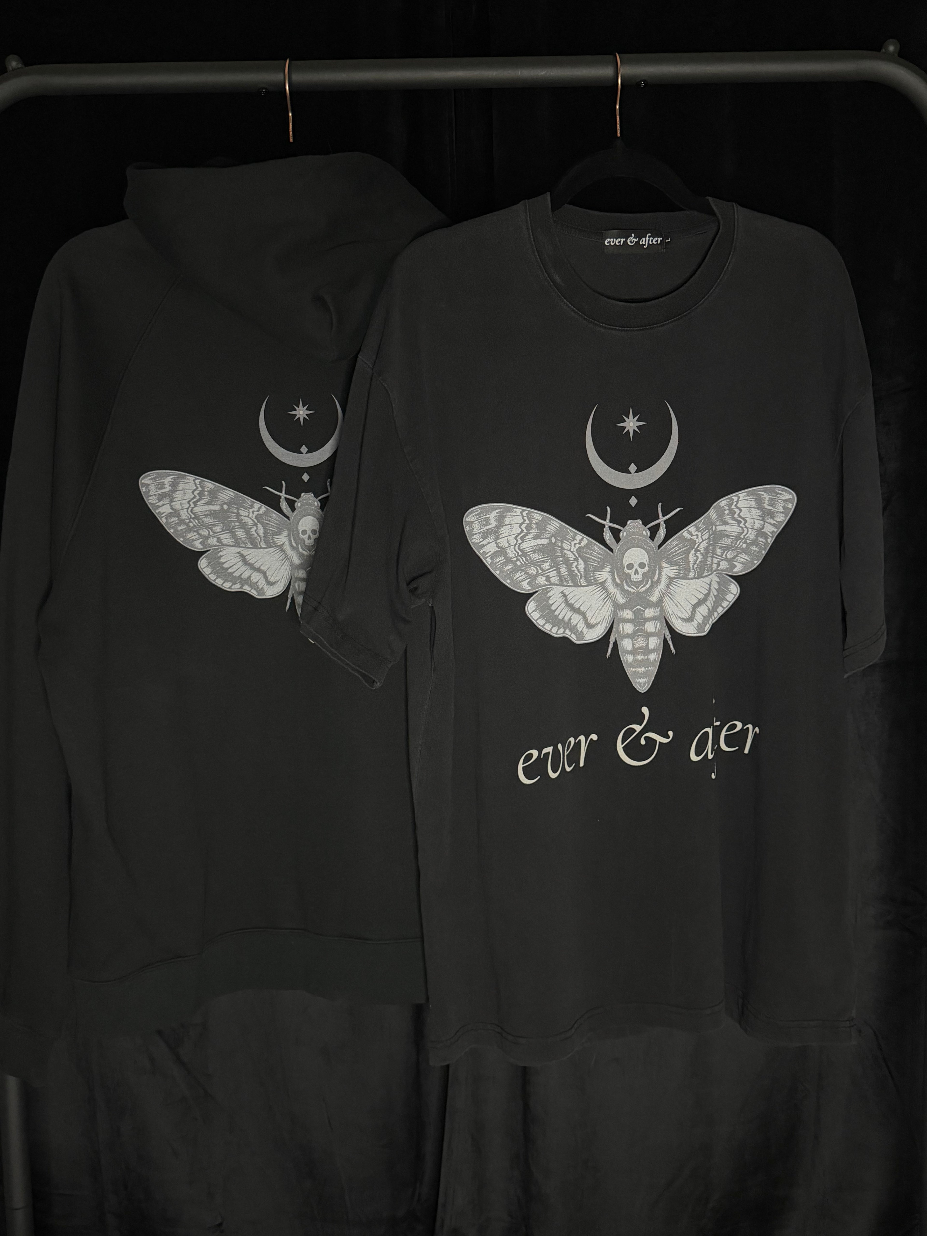 A black hoodie and matching black t-shirt hanging on a rack against a dark background. Both garments feature a large white moth graphic with a crescent moon and star above it, with the t-shirt also displaying the text 'ever & aer' below the graphic.