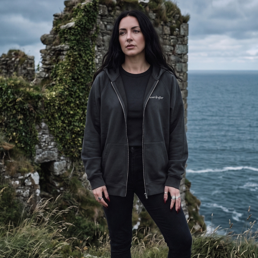 Woman wearing a dark jacket with text standing in front of a castle ruin and ocean.