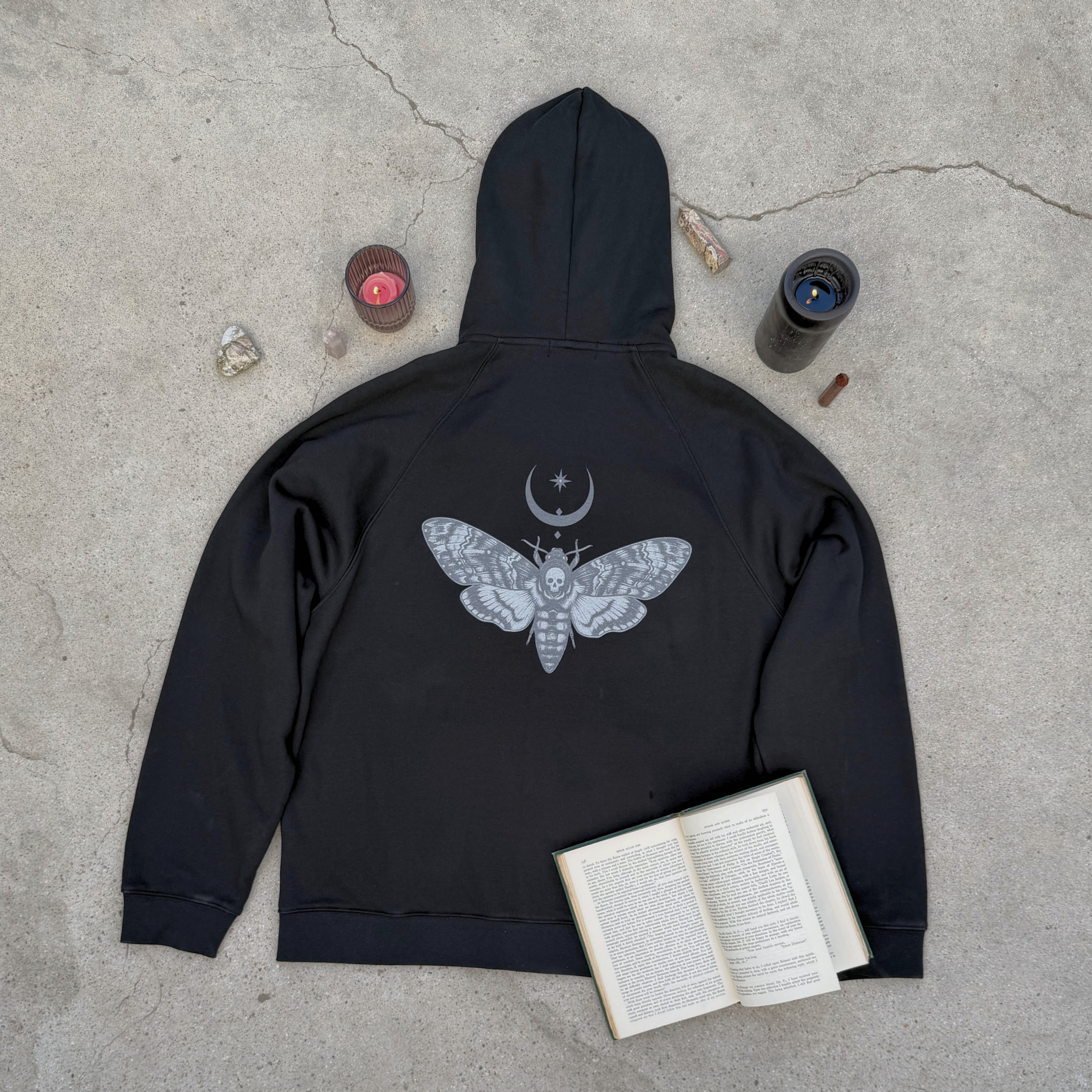 Black hoodie with moth design on a concrete surface with books and candles.