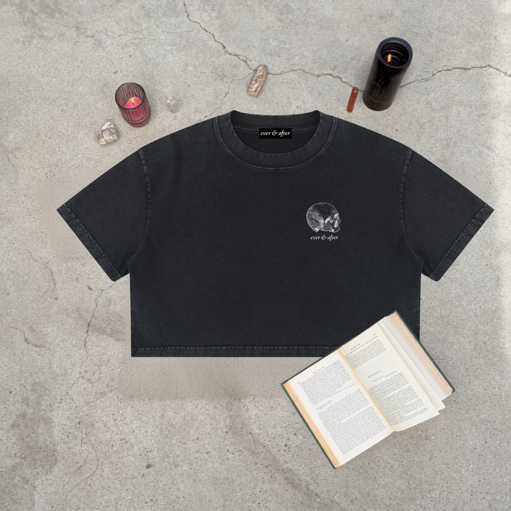 A black cropped t-shirt with a small white skull graphic and 'run of fate' text on the left chest, styled in a flat-lay on a concrete surface with an open book, crystals, and candles.