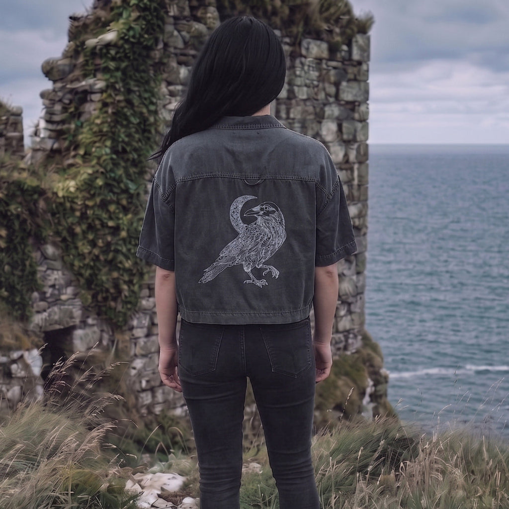 Person wearing a dark jacket with a bird design in front of stone ruins and ocean.