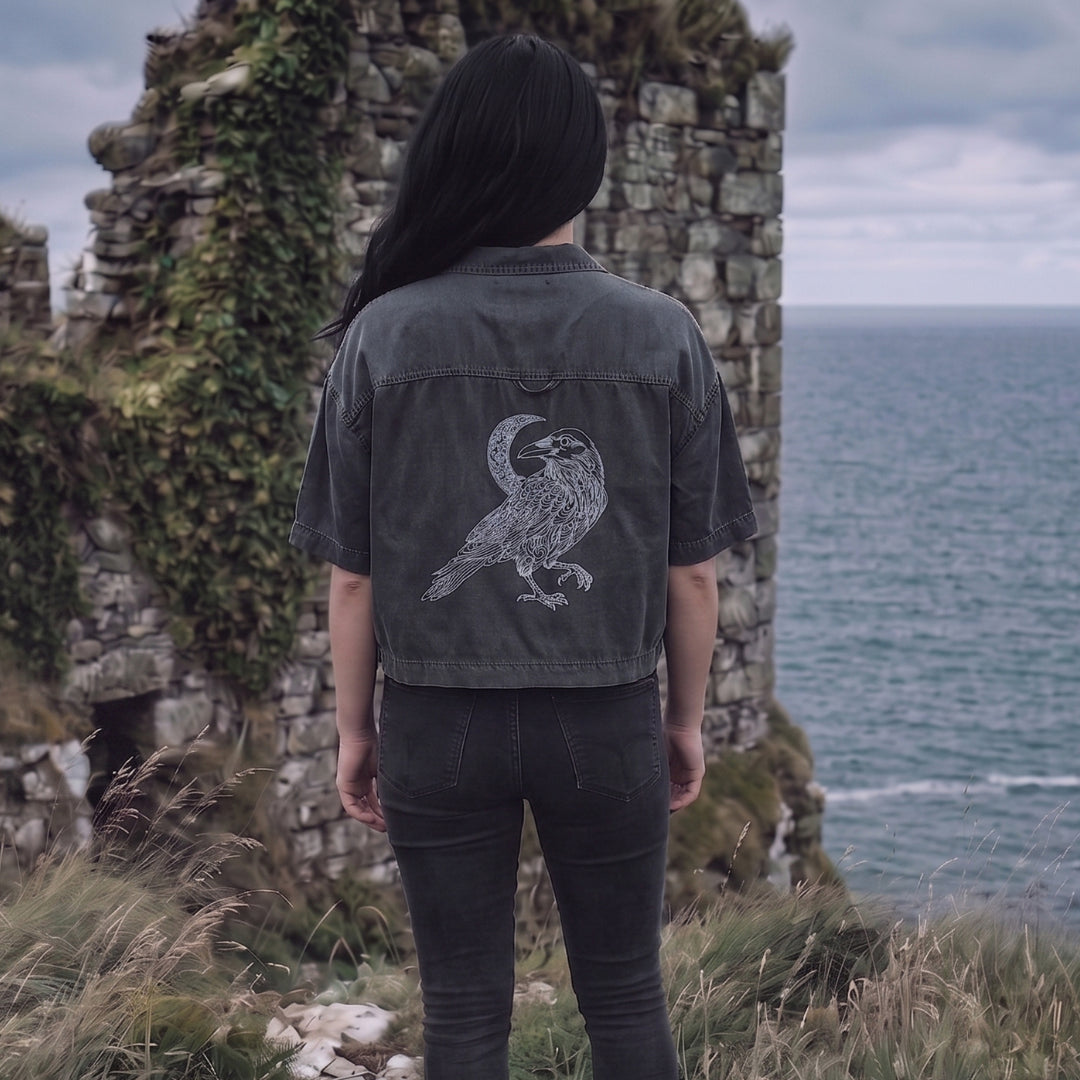 Person wearing a dark jacket with a bird design in front of stone ruins and ocean.
