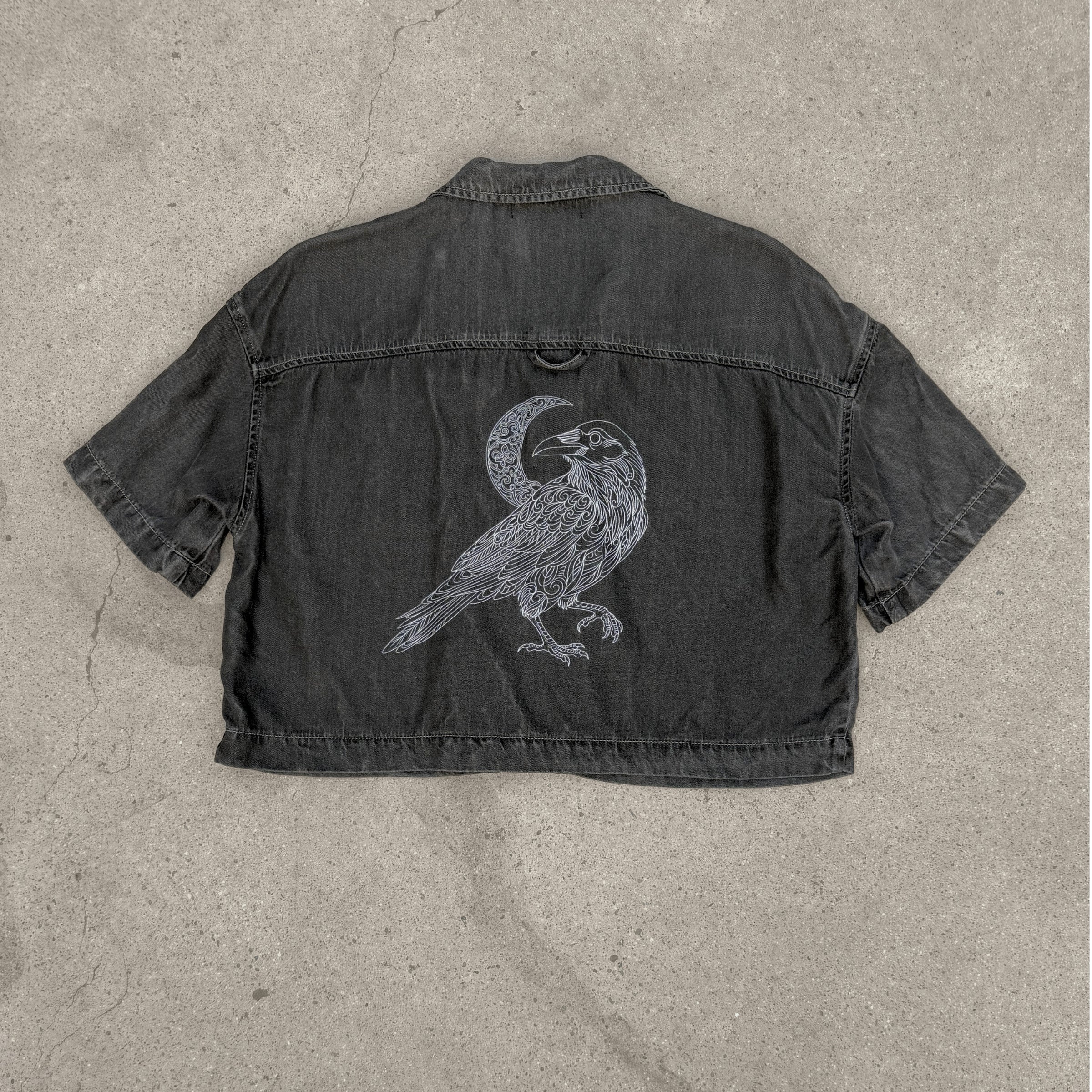Black denim jacket with a bird and moon design on a concrete surface